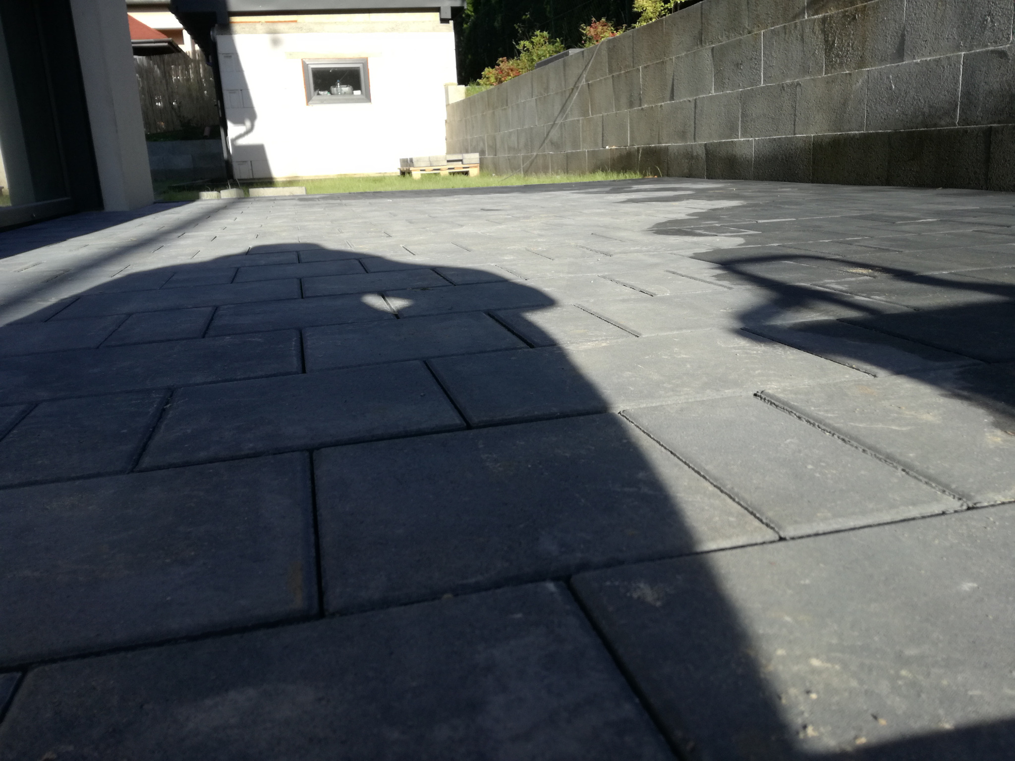 Ułożona szara kostka brukowa na podwórku z widocznym cieniem osoby fotografującej, w tle budynek z białymi ścianami i fragment muru z szarych kamieni.