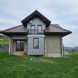 Remkop - Dwupiętrowy dom jednorodzinny z szarą elewacją z kamienia, brązowym dachem z dachówki, balkonem z ozdobną balustradą, widoczne drabiny i akcesoria budowlane, na tle zielonego wzgórza.