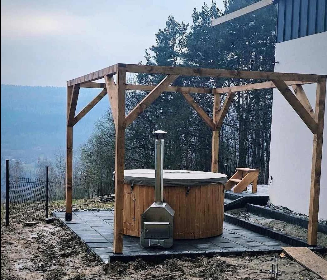 Drewniana balia z piecem na zewnątrz, zadaszona drewnianą konstrukcją, na wyłożonym płytkami tarasie, z widokiem na zalesione wzgórza.