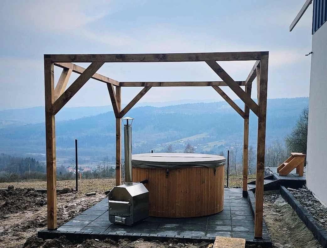 Drewniana balia z piecem na zewnątrz, zadaszona drewnianą konstrukcją, na tarasie z szarych płyt, w tle górzysty krajobraz.