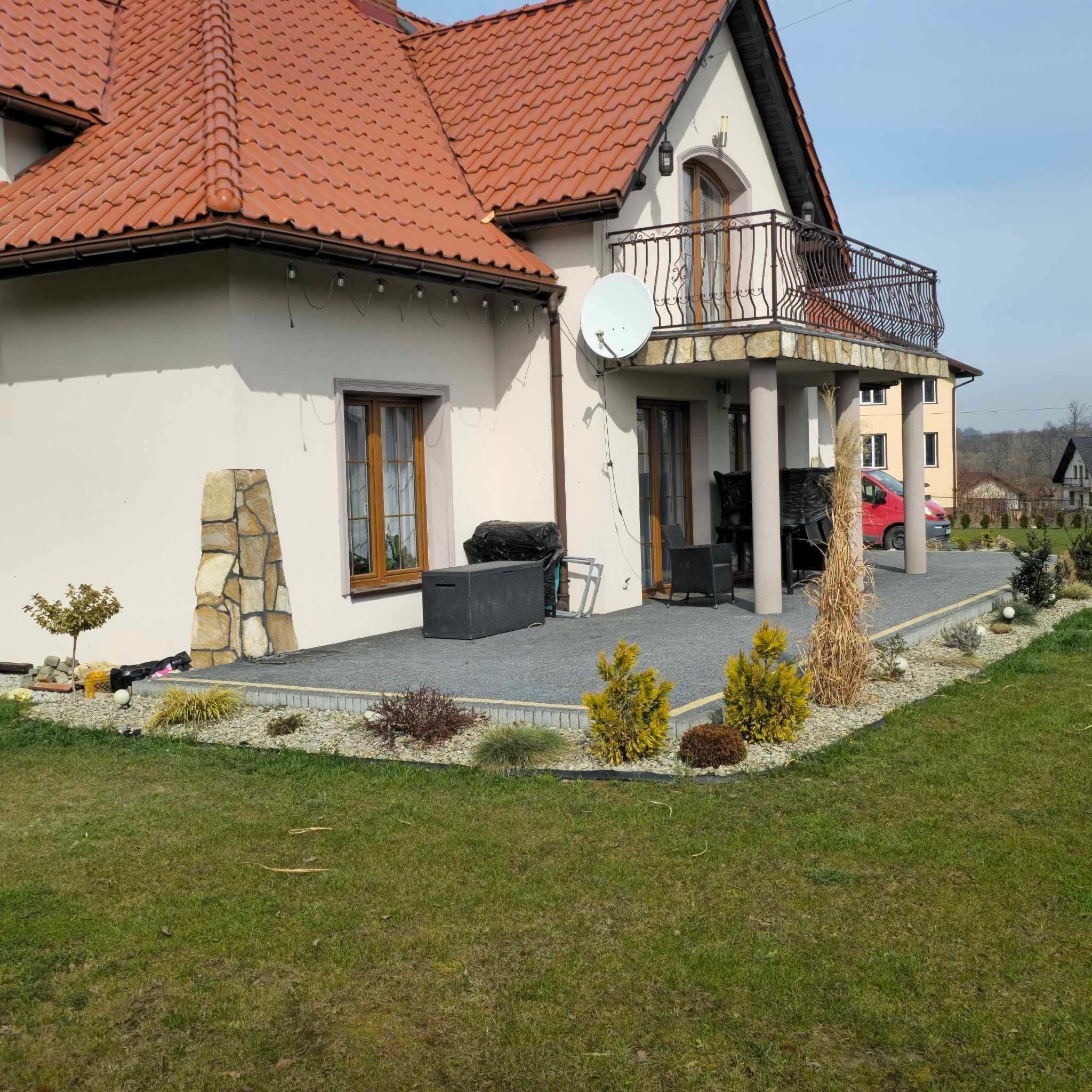 Dom z tarasem wyłożonym szarą kostką, fragment elewacji z kamienia dekoracyjnego, ogródek z ozdobnymi krzewami i trawnikiem.