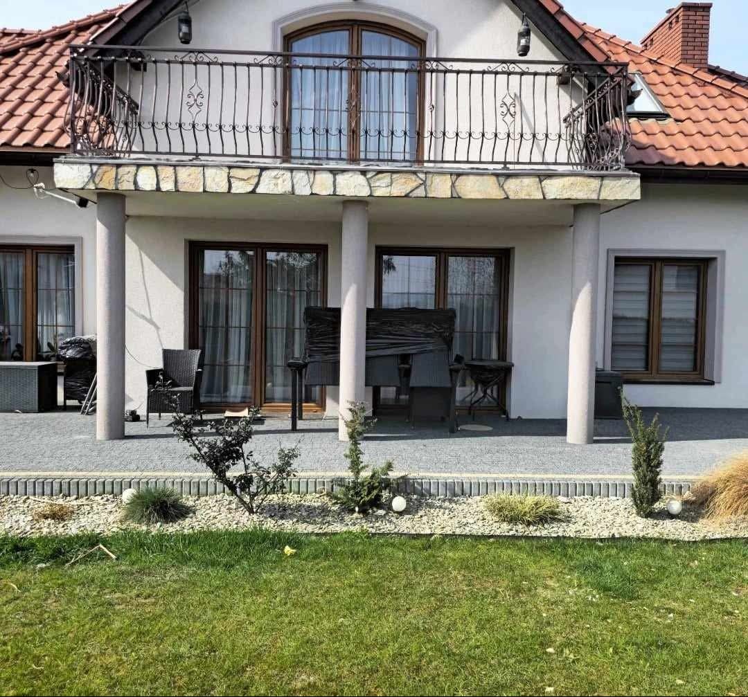Elewacja domu z balkonem i tarasem, część mebli na tarasie zabezpieczona folią, widoczny trawnik i elementy dekoracyjne ogrodu.