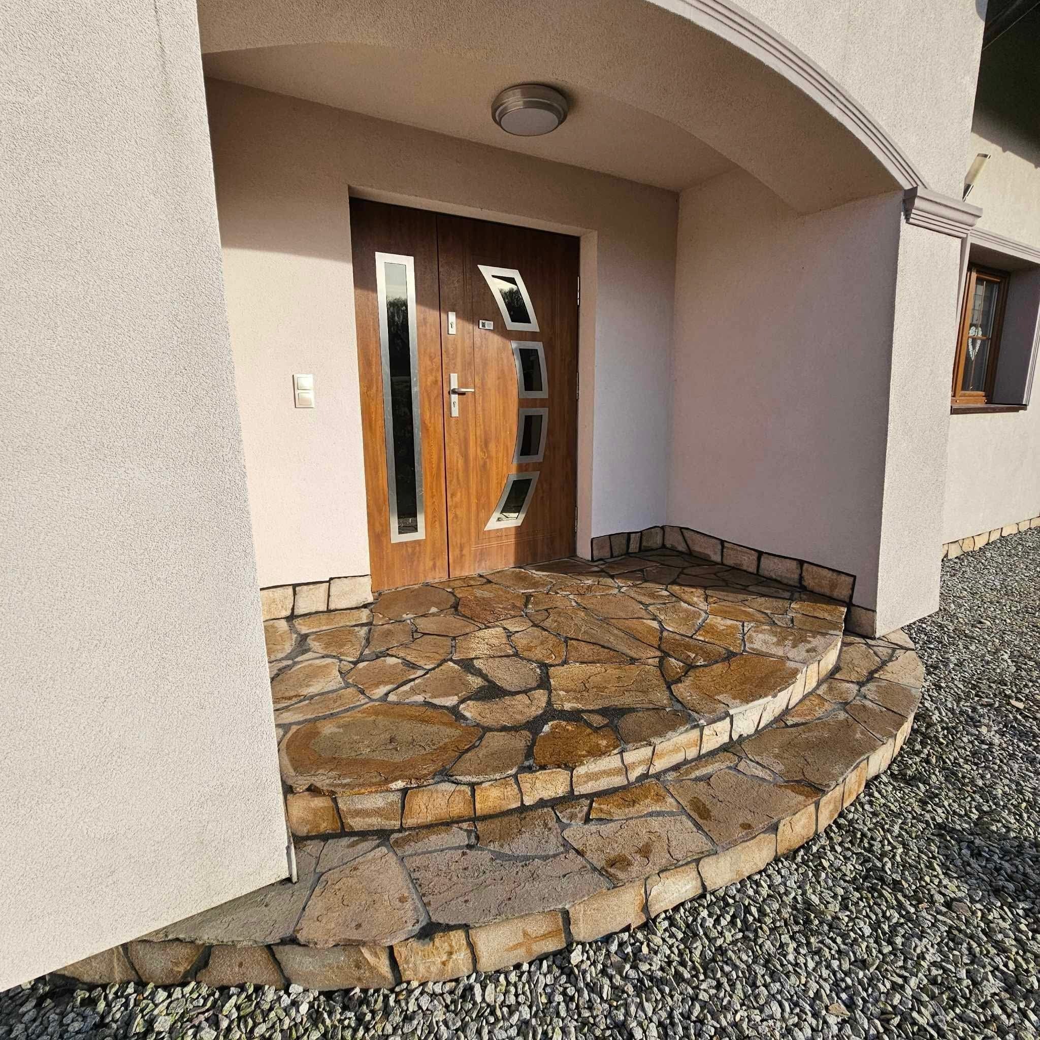 Wejście do domu z dwustopniowymi schodami wyłożonymi nieregularnymi kamiennymi płytami w odcieniach brązu i beżu, brązowe drzwi z przeszkleniami, ściany w jasnym odcieniu beżu, oświetlenie...