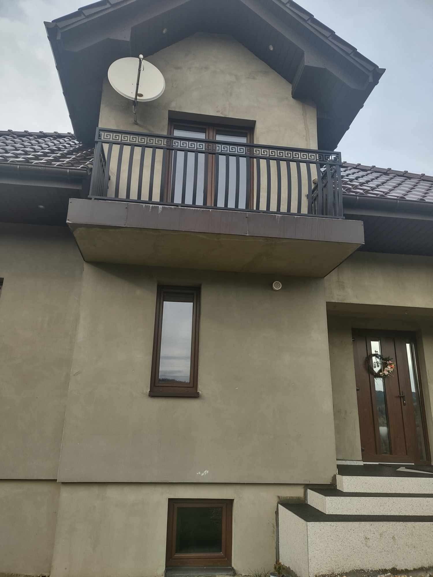 Dwupiętrowy dom z balkonem z czarną balustradą z motywem greckim, brązowymi oknami i drzwiami wejściowymi ozdobionymi wieńcem, na ścianie satelita.