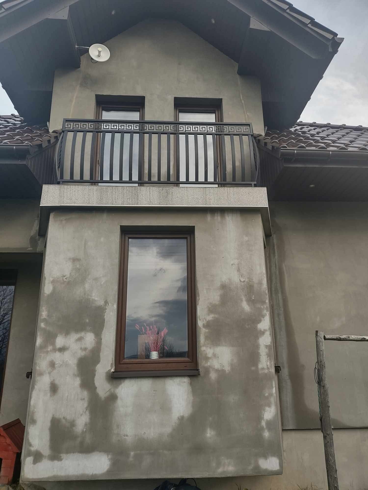 Surowa elewacja domu z balkonem z metalową balustradą o greckim wzorze, widoczne ślady po pracach murarskich i okno z odbijającym się niebem.
