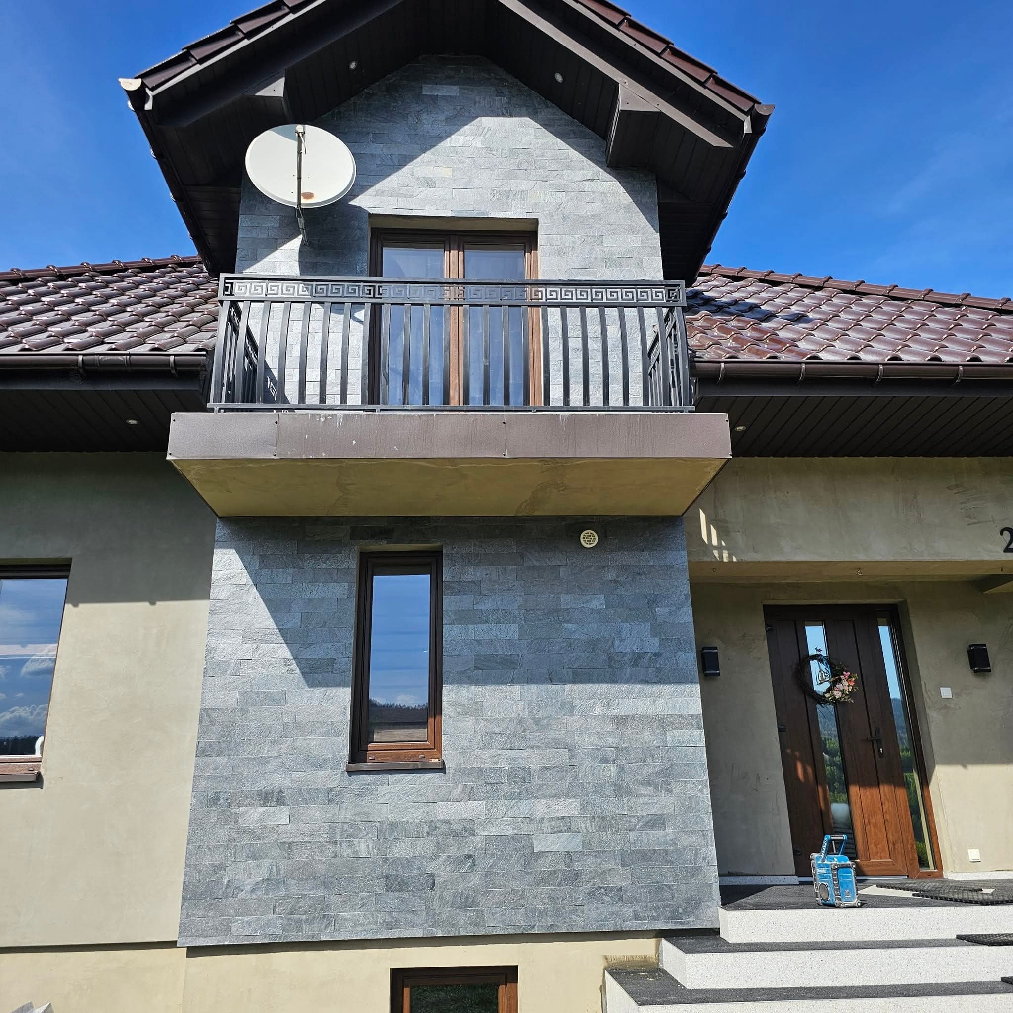 Elewacja domu z szarej cegły, balkon z metalową balustradą, brązowe okna i dachówka, antena satelitarna, wejście z dekoracyjnym wieńcem i turkusowym pojemnikiem na schodach.