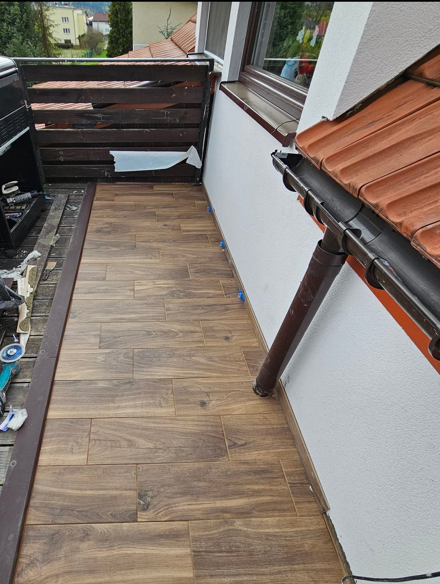 Balkon z nową, drewnopodobną podłogą z płytek, widoczne narzędzia i akcesoria montażowe, fragment zadaszenia i rynny.