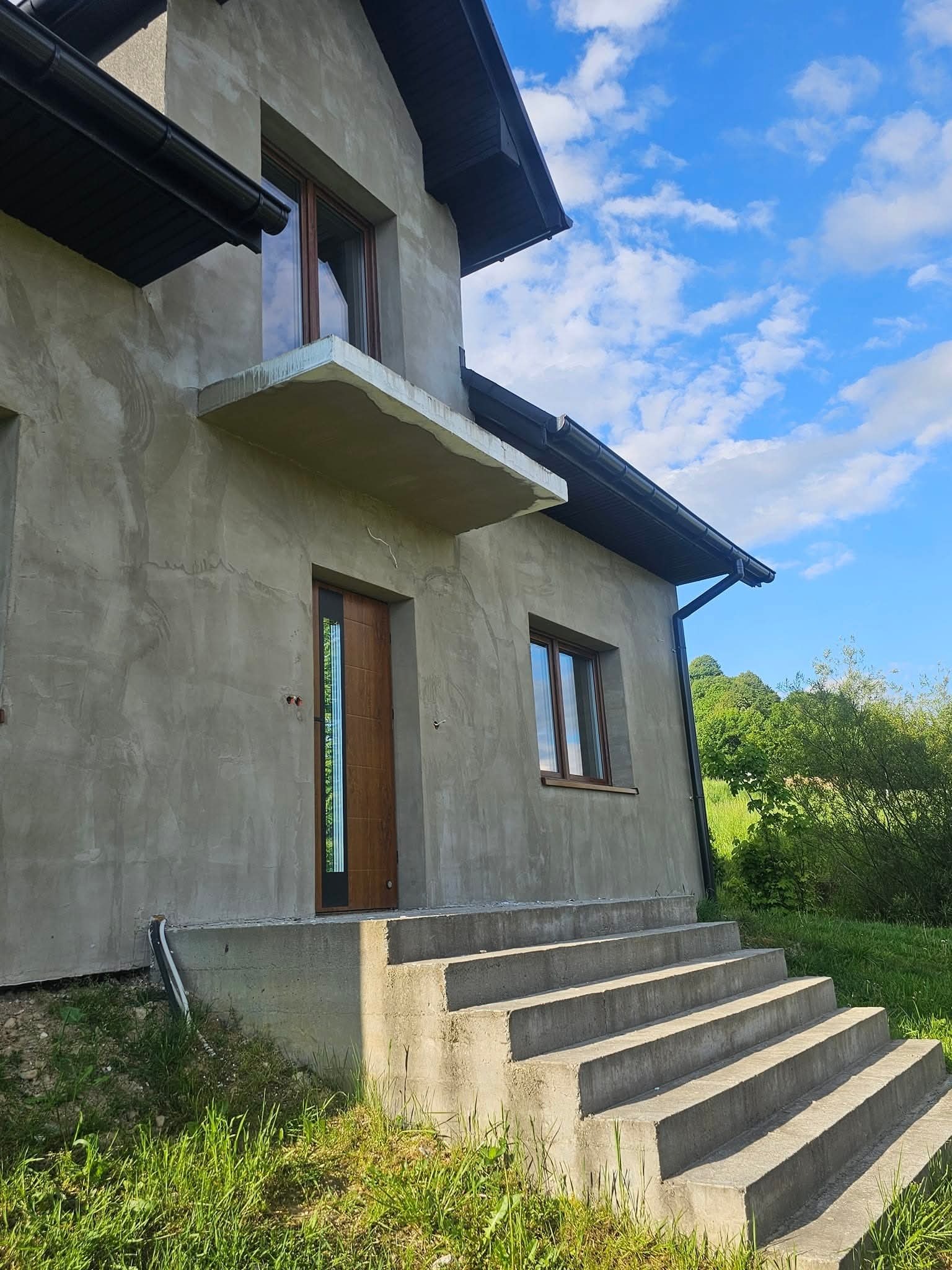 Surowy, otynkowany dom jednorodzinny z drewnianymi oknami i drzwiami, betonowymi schodami wejściowymi, widok w słoneczny dzień.