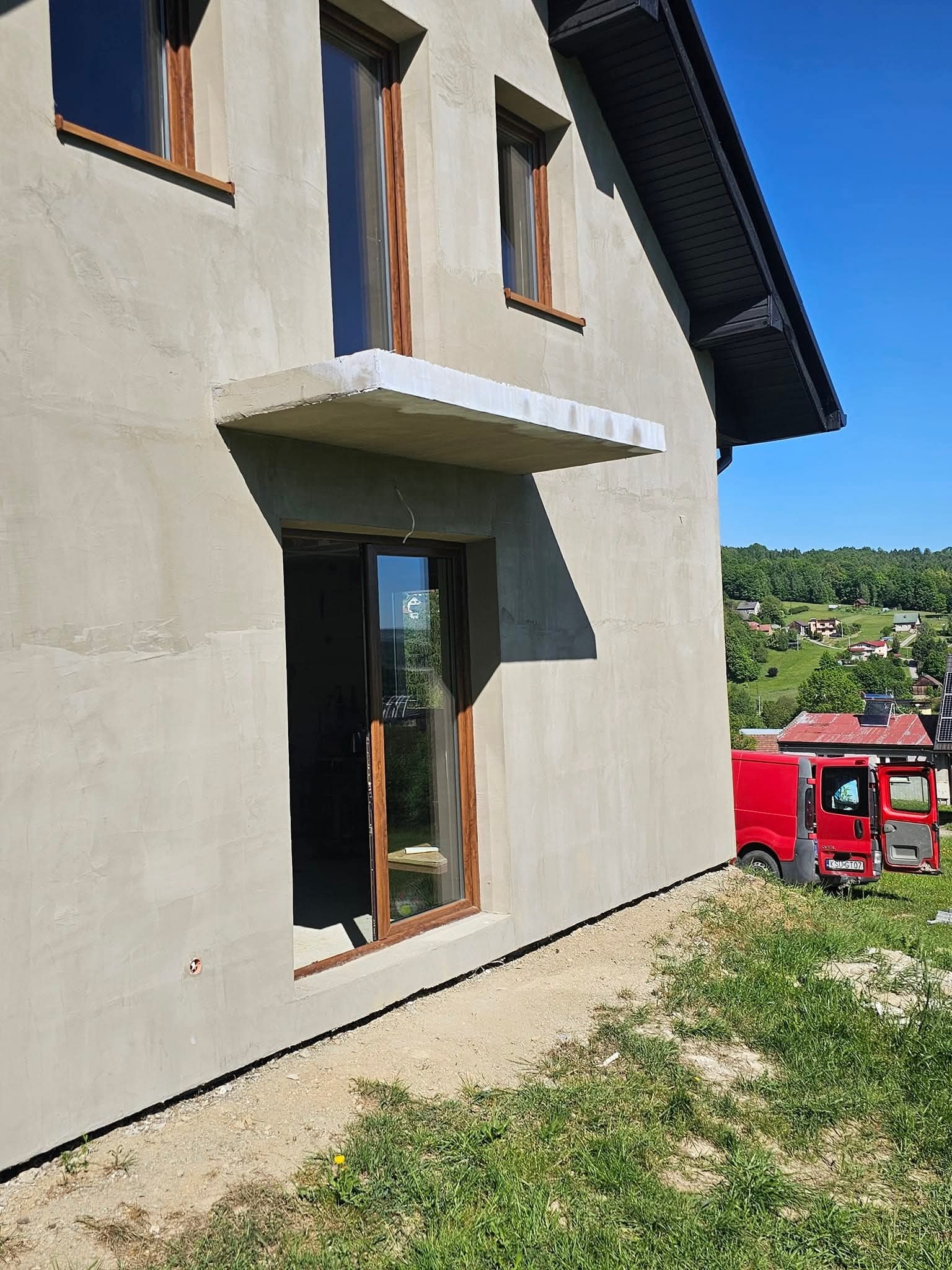 Surowa elewacja domu w trakcie prac, widoczne okna z brązowymi ramami, betonowy daszek nad oknem, czerwony samochód dostawczy w tle, teren wokół domu jeszcze niezagospodarowany.