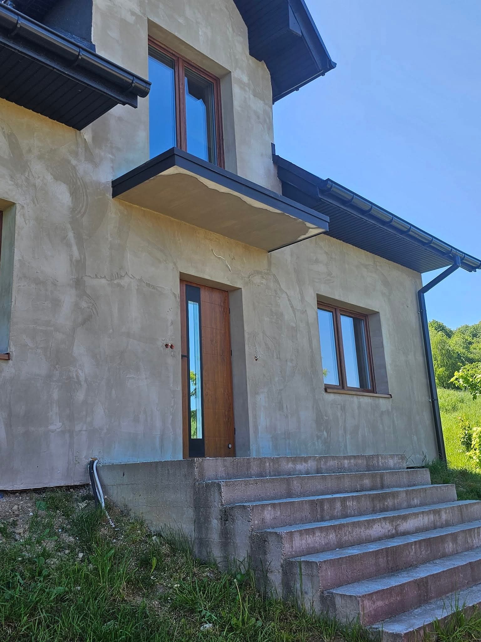 Surowa elewacja domu jednorodzinnego z drewnianą stolarką okienną i drzwiami wejściowymi, betonowe schody prowadzące do drzwi, widoczne orynnowanie i fragment zieleni w tle.