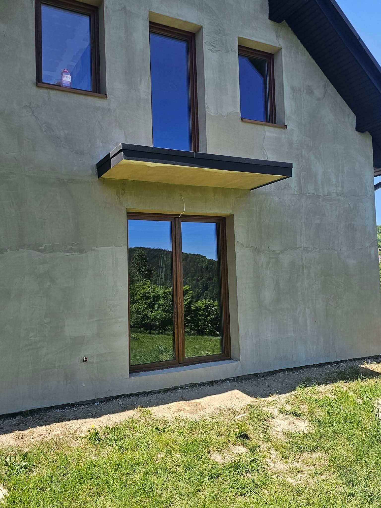 Surowy tynk na elewacji domu jednorodzinnego z drewnianą stolarką okienną, widoczny fragment zadaszenia nad oknem i butelka wody na parapecie w górnym oknie.