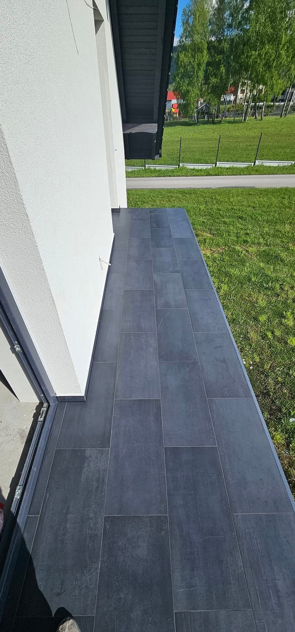 Nowoczesny taras wyłożony ciemnoszarymi płytkami ceramicznymi, widok z boku budynku na zielony trawnik i ogrodzenie.