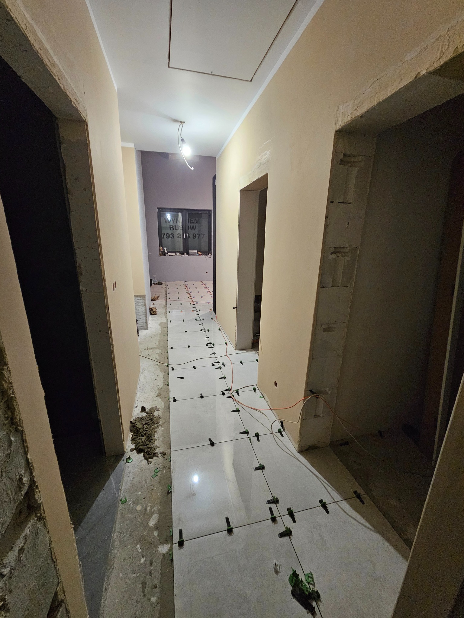 Korytarz w trakcie układania płytek podłogowych, widoczne krzyżyki dystansowe, ściany w surowym stanie, w tle okno z naklejonym numerem telefonu.