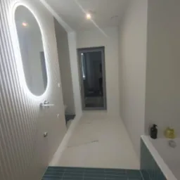 Nowoczesna łazienka z podświetlanym lustrem o owalnym kształcie, białymi ścianami z pionowymi żłobieniami i turkusową mozaiką podłogową, widok na przeszklone drzwi w głębi.