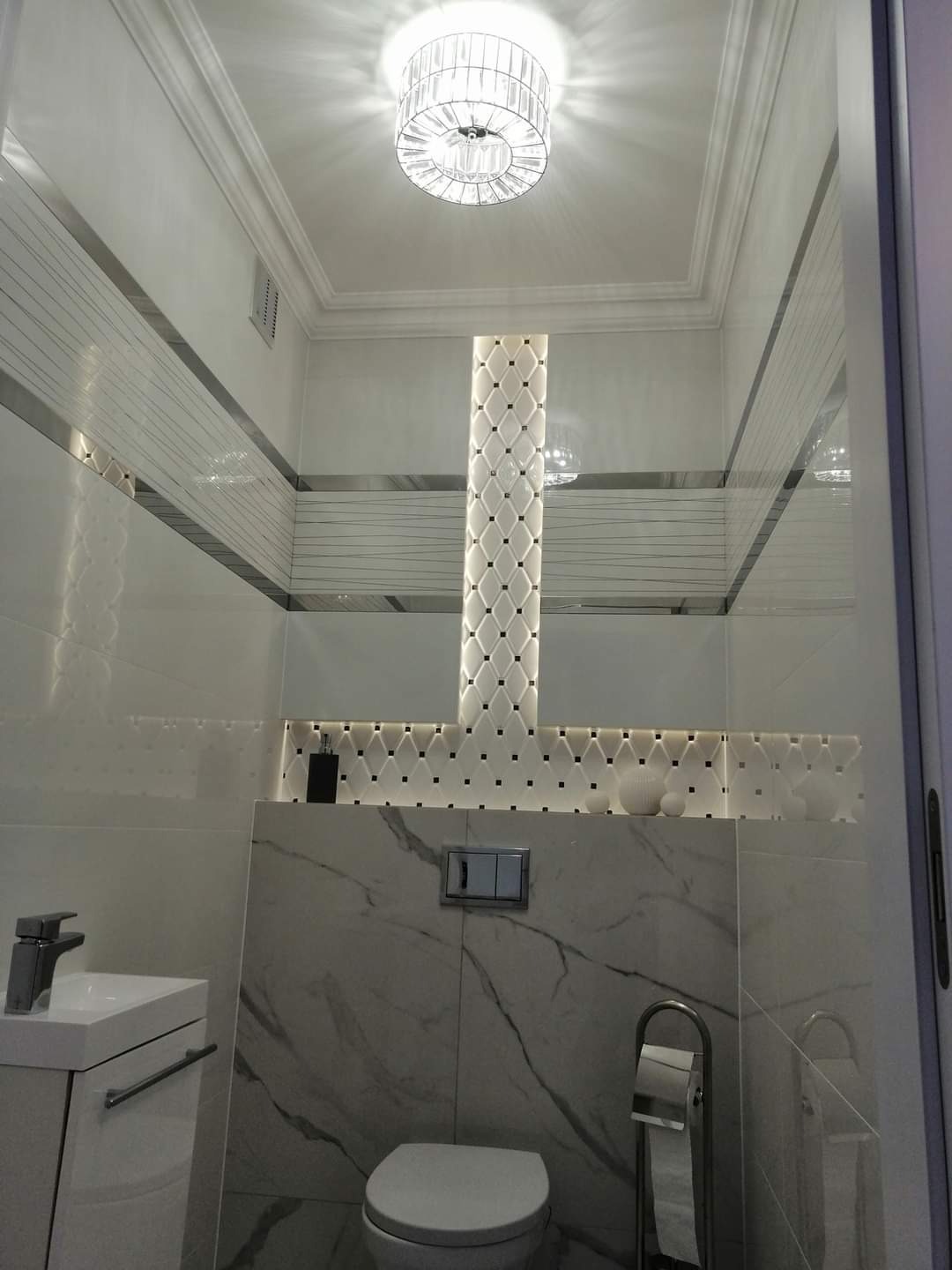 Elegancka, biało-szara łazienka z podświetlaną kolumną z płytek dekoracyjnych, marmurowymi płytkami na ścianach i nowoczesną armaturą, oświetlona kryształowym żyrandolem.