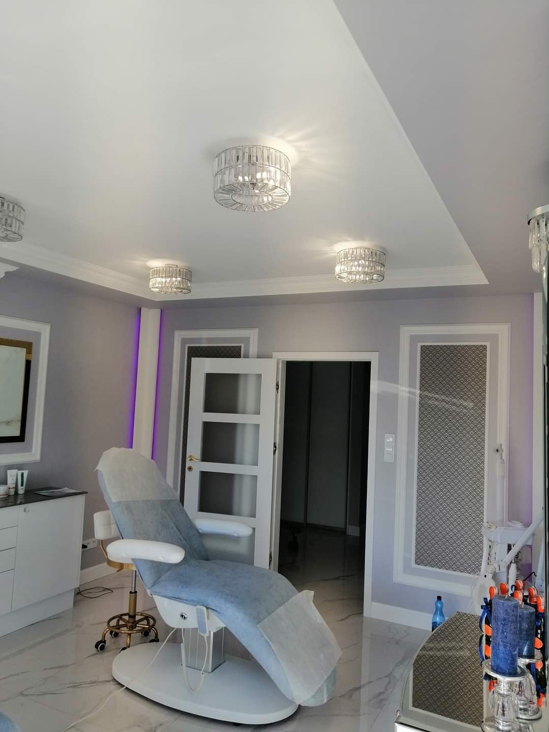 Jasne wnętrze gabinetu kosmetycznego z marmurową podłogą, szarymi ścianami, trzema kryształowymi lampami sufitowymi i fotelem kosmetycznym z niebieskim obiciem zabezpieczonym jednorazową podkładką.