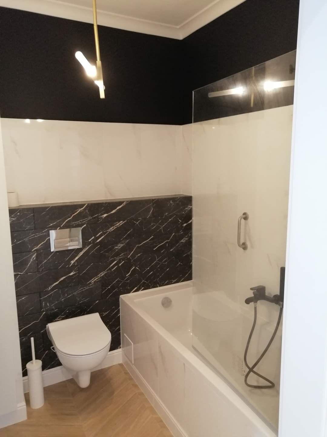 Nowoczesna łazienka z czarnym sufitem i marmurowymi płytkami, minimalistyczna toaleta podwieszana, szklana kabina nawannowa z czarną armaturą, drewnopodobna podłoga ułożona w jodełkę.