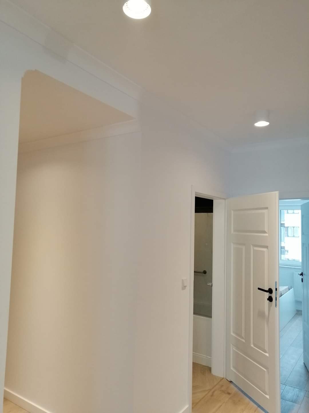 Wnętrze po odświeżeniu: białe ściany z listwami przypodłogowymi i sufitowymi, drewniana podłoga, otwarte drzwi do łazienki z widoczną wanną i do pokoju z jasnymi meblami.