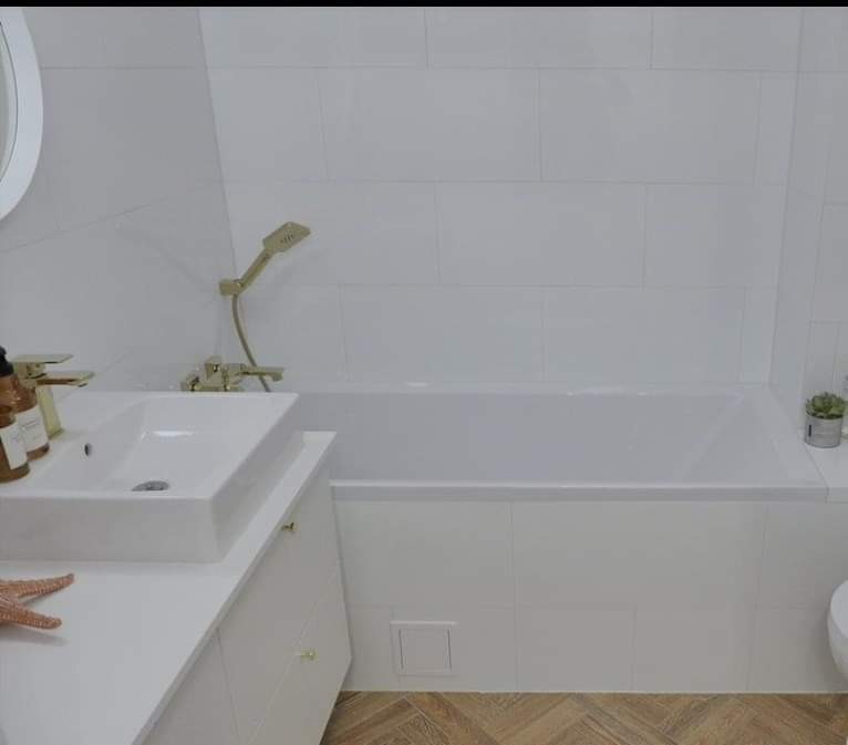 Nowoczesna biała łazienka z wanną obłożoną kafelkami, umywalką nablatową, złotą armaturą i drewnopodobną podłogą w jodełkę.