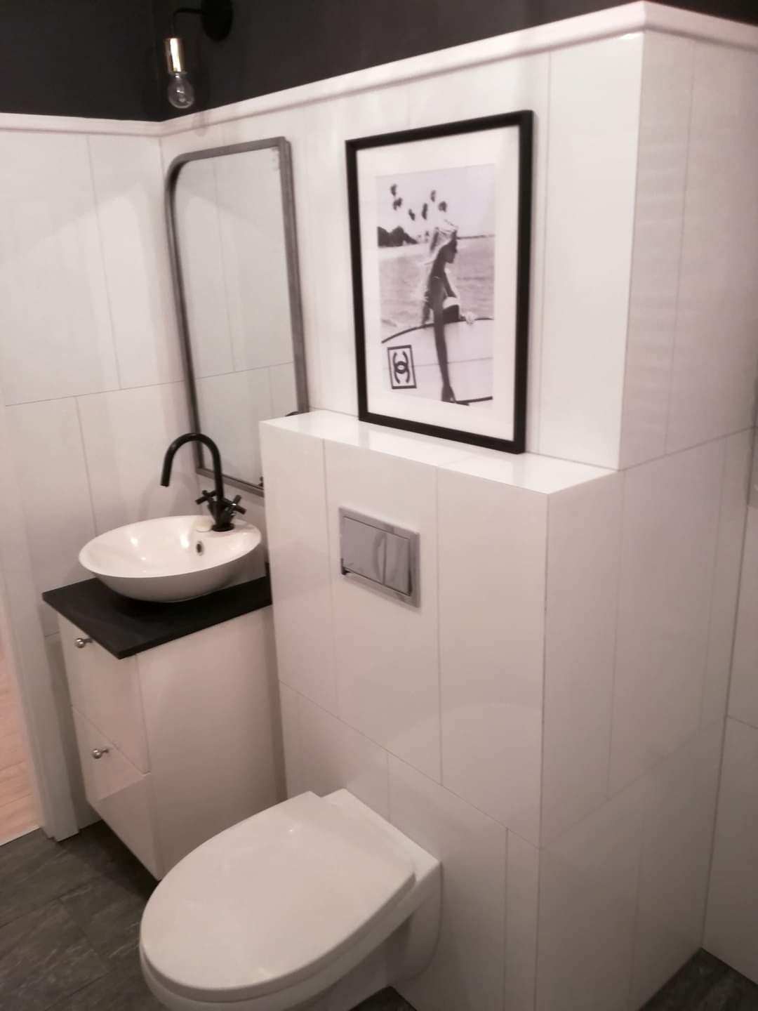 Nowoczesna łazienka z białą armaturą, umywalką nablatową na szafce z czarnym blatem, wiszącą toaletą i czarno-białym zdjęciem w ramie na ścianie pokrytej białymi płytkami.