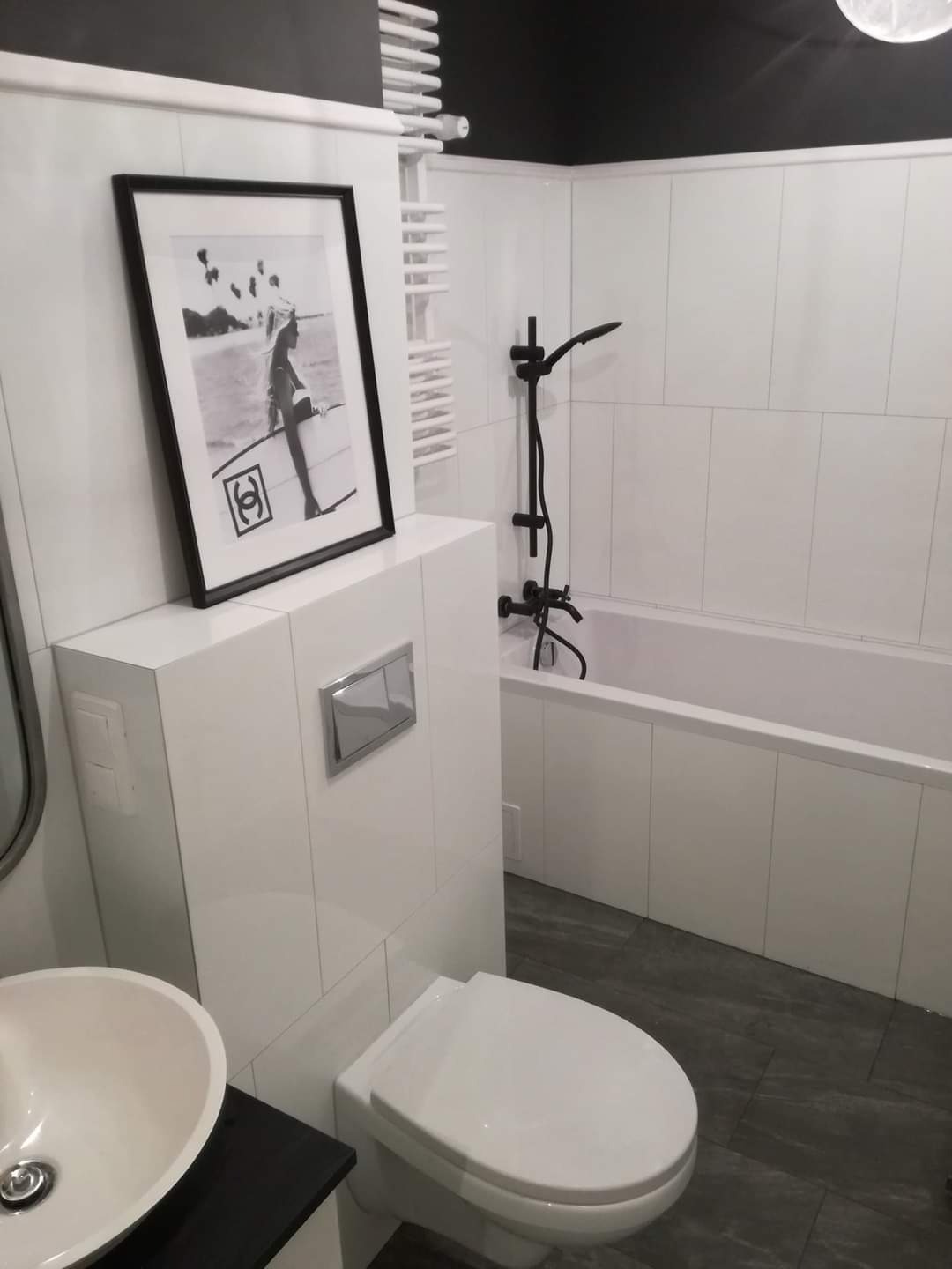 Nowoczesna łazienka z białymi płytkami ściennymi, czarną armaturą, podwieszaną toaletą i czarno-białym plakatem w ramie nad nią.