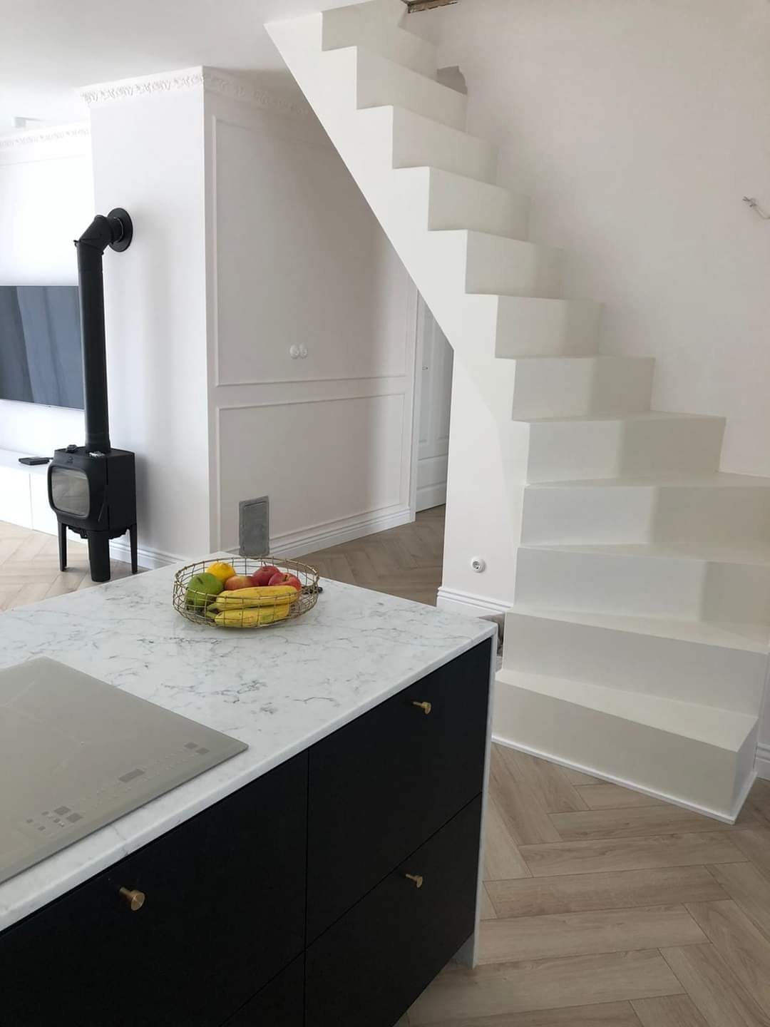 Nowoczesna kuchnia z czarnymi szafkami, marmurowym blatem, owocami w drucianym koszyku, piecem wolnostojącym i białymi schodami bez poręczy w tle.