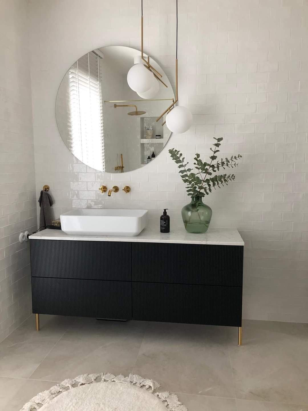 Nowoczesna łazienka z czarną szafką pod umywalkę, białym blatem i okrągłym lustrem; złote akcenty w postaci baterii, nóżek szafki i lampy wiszącej; białe płytki na ścianach i jasna podłoga...