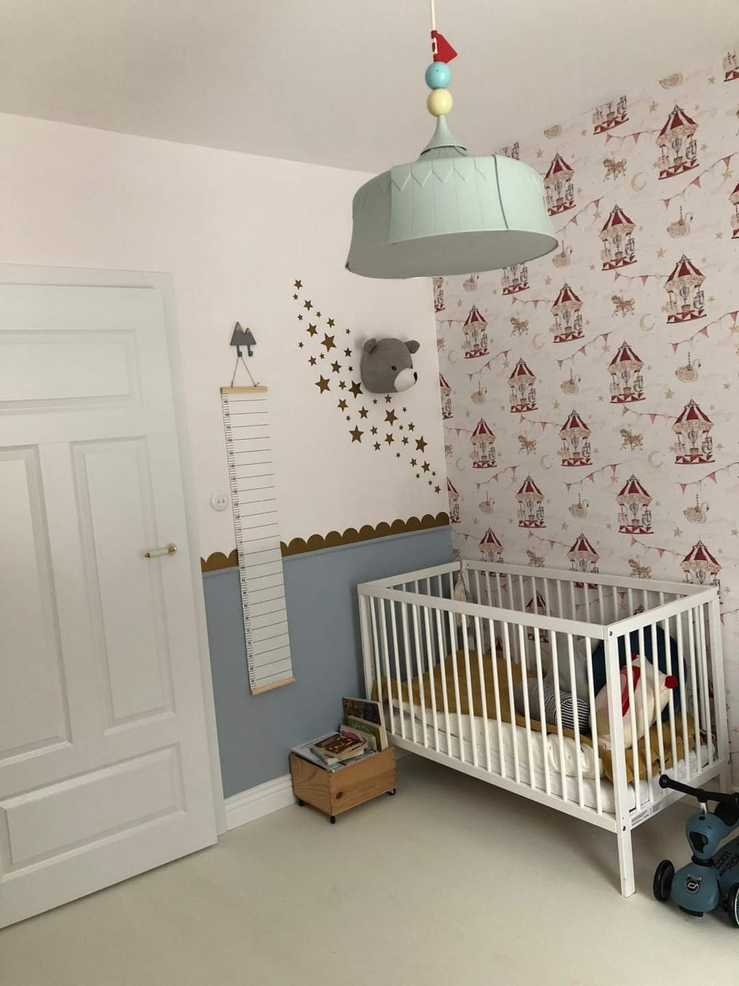 Umeblowany pokój dziecięcy z łóżeczkiem, lampą sufitową w kształcie karuzeli, tapetą z motywem cyrkowym i dekoracjami ściennymi w postaci gwiazdek i pluszowego misia.