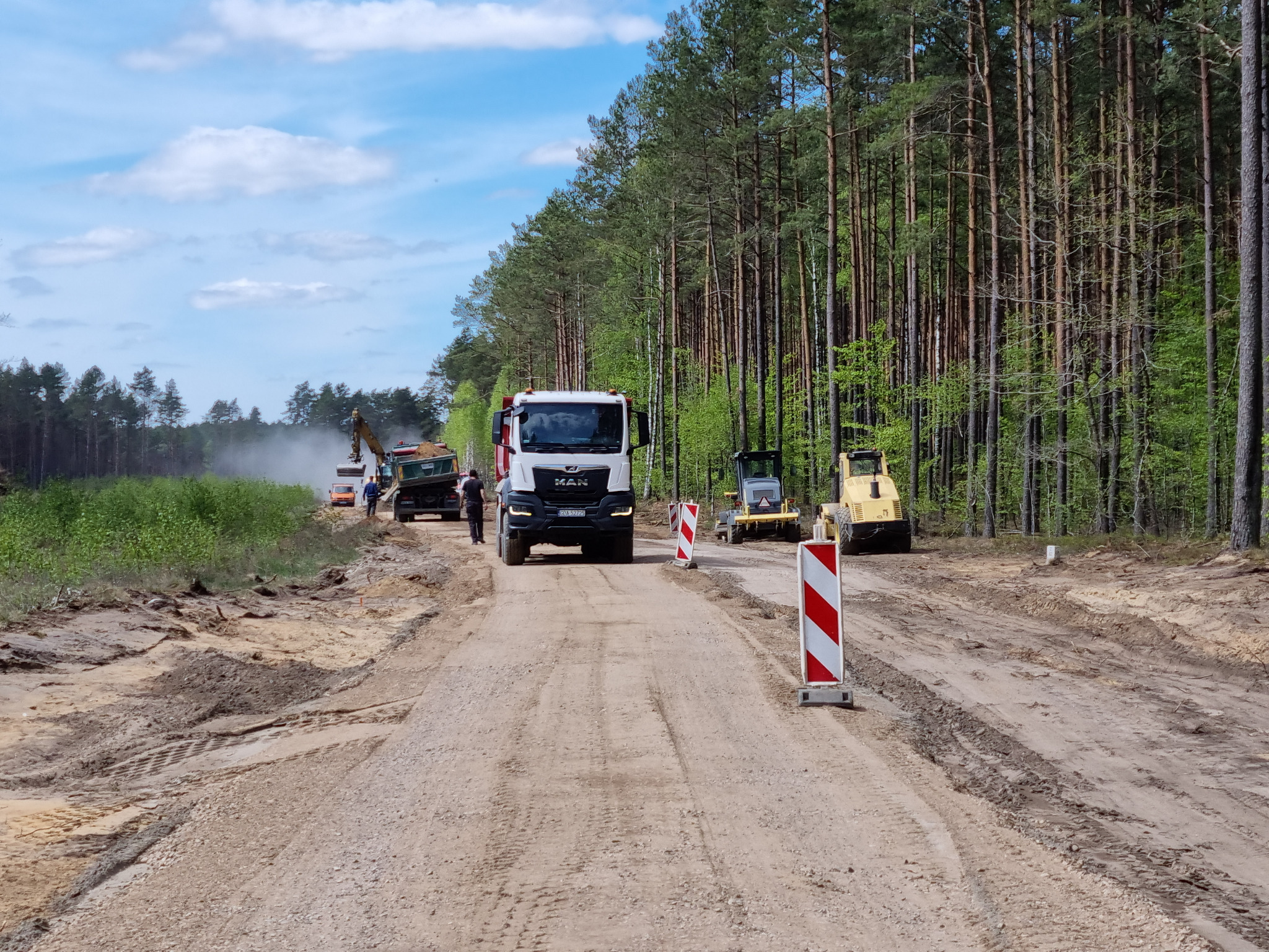 Ciężarówka MAN na drodze gruntowej w trakcie prac ziemnych, w tle las i maszyny budowlane, widoczne słupki ostrzegawcze.
