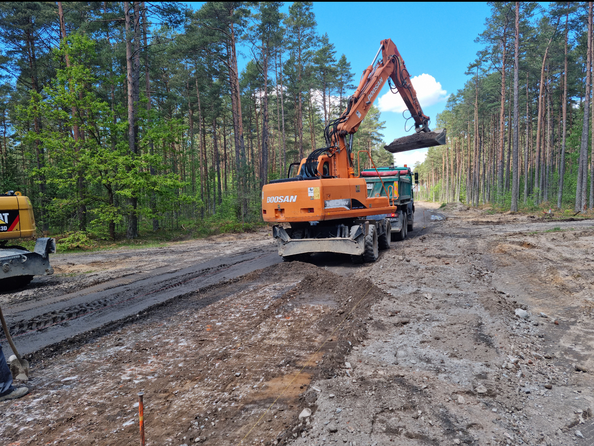 Pomarańczowa koparka Doosan ładuje ziemię do zielonej wywrotki na tle lasu sosnowego, widoczny częściowo żółty walec i przygotowany teren pod drogę.
