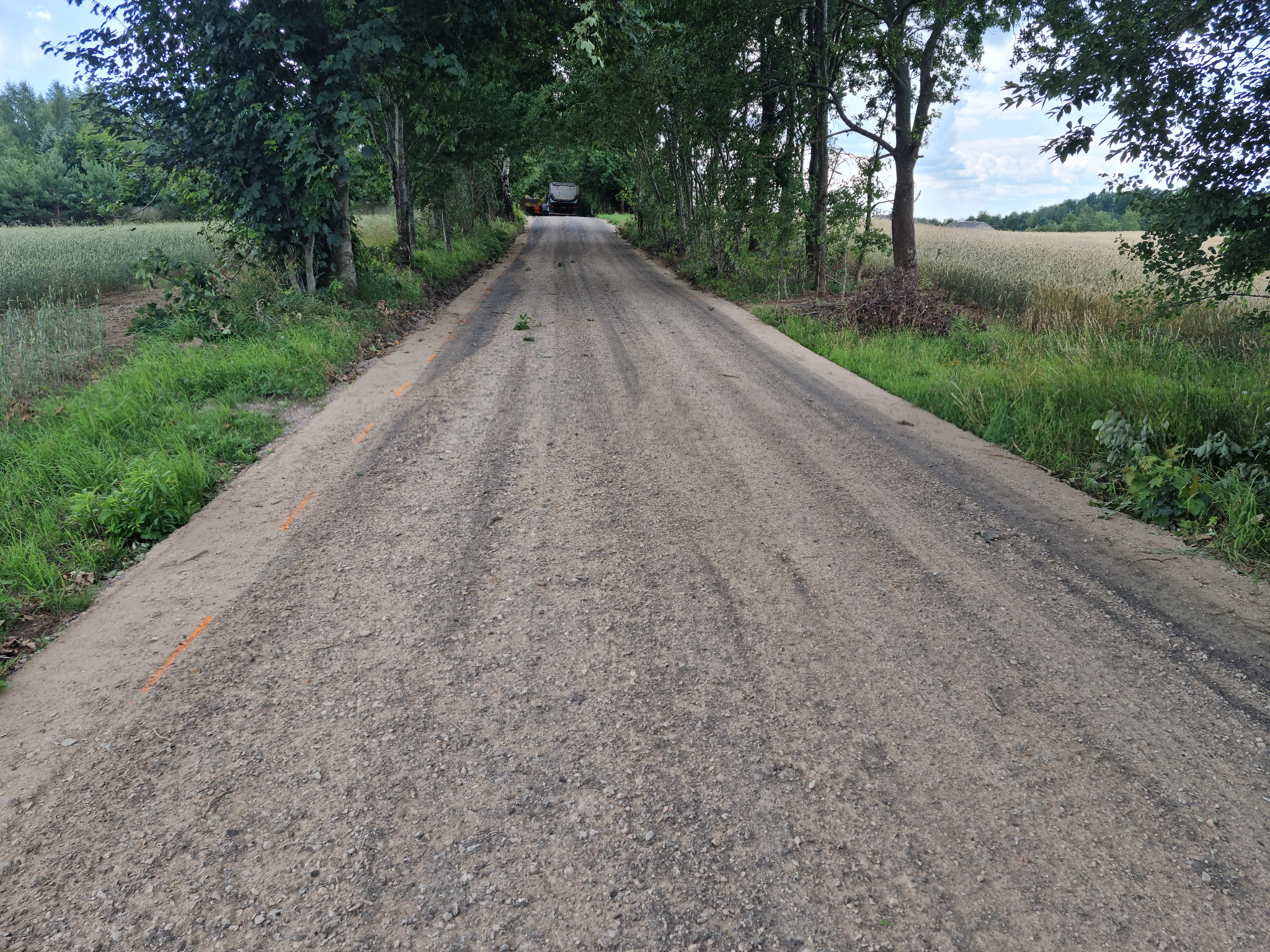 Polna droga gruntowa z widocznymi śladami opon, otoczona drzewami i polami, z odległym widokiem na maszynę budowlaną.