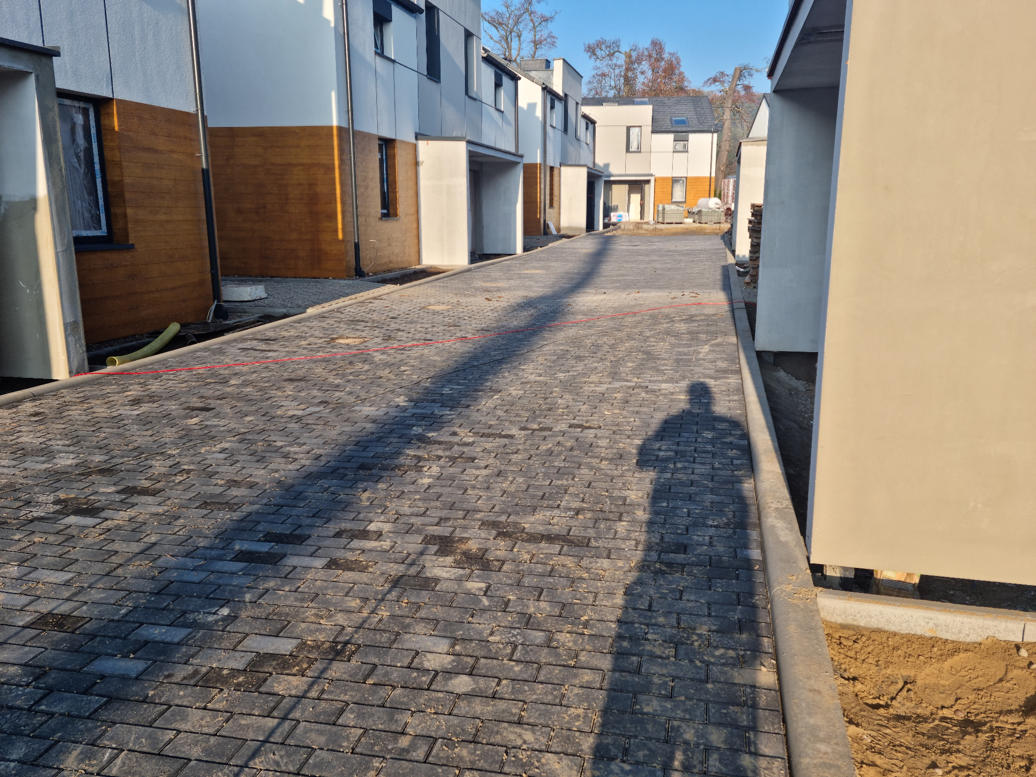 Nowa, ciemnoszara kostka brukowa ułożona na osiedlu domów jednorodzinnych w zabudowie szeregowej, widoczny cień osoby fotografującej.