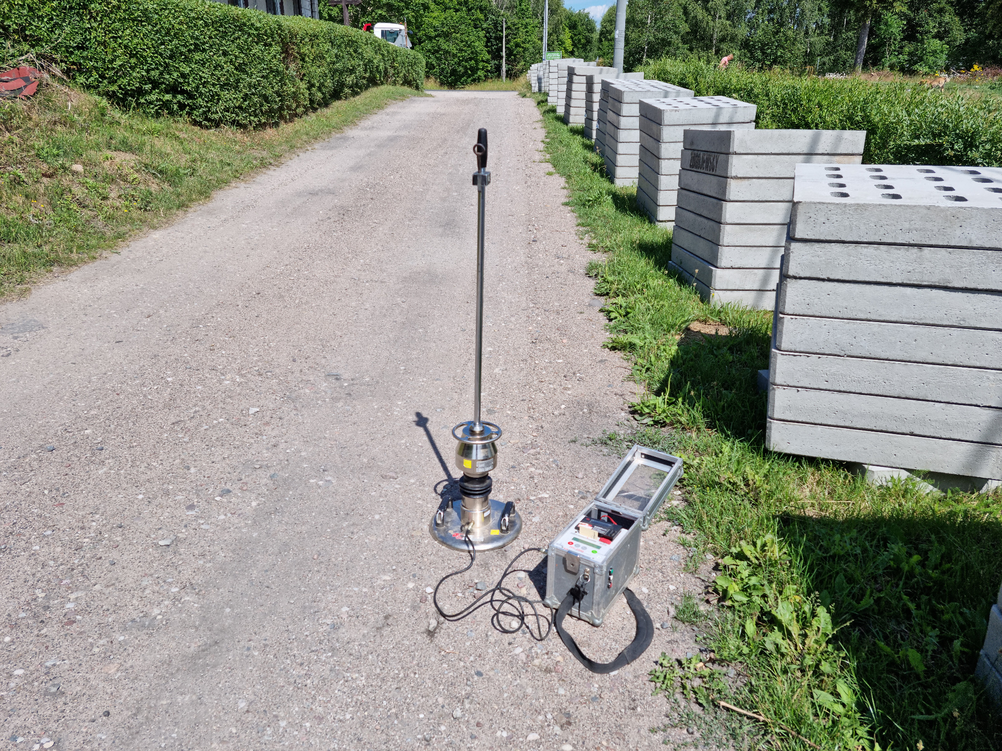 Badanie zagęszczenia gruntu w Brodnicy Górnej za pomocą dynamicznego sondowania płytą obciążaną, obok składowane betonowe prefabrykaty z otworami.