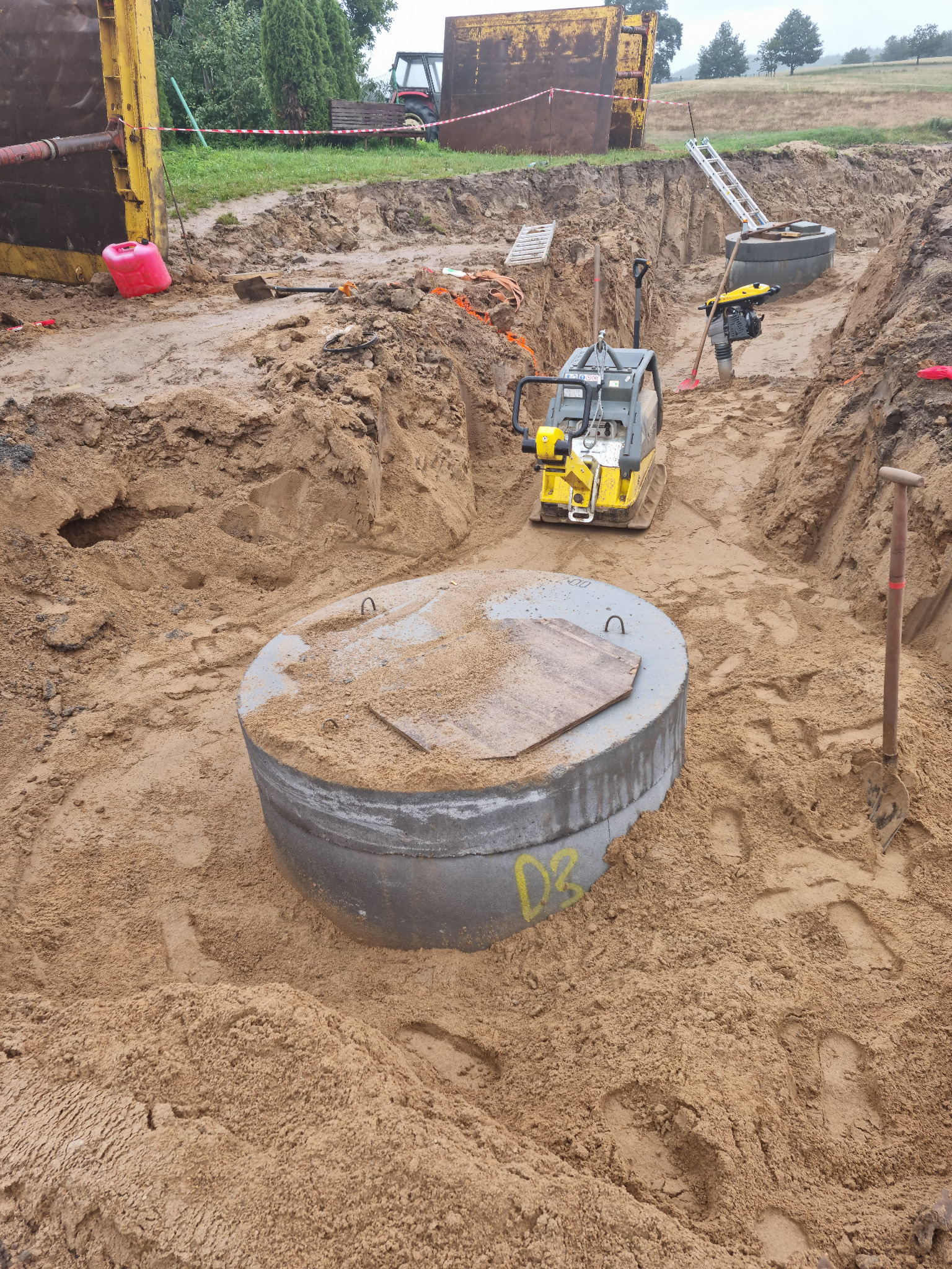 Wykop z betonowym kręgiem studziennym w piasku, ubijarka wibracyjna i szalunki widoczne w tle.