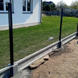 Wykonywanie fundamentu pod ogrodzenie z prefabrykowanych elementów betonowych, widoczne czarne słupki, w tle trawnik i budynek.