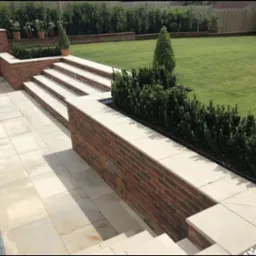 Ogród z tarasem i schodami wyłożonymi jasną kostką brukową, ceglana ścianka z wbudowanymi rabatami, trawnik z równo przyciętą trawą i żywopłotem w tle.