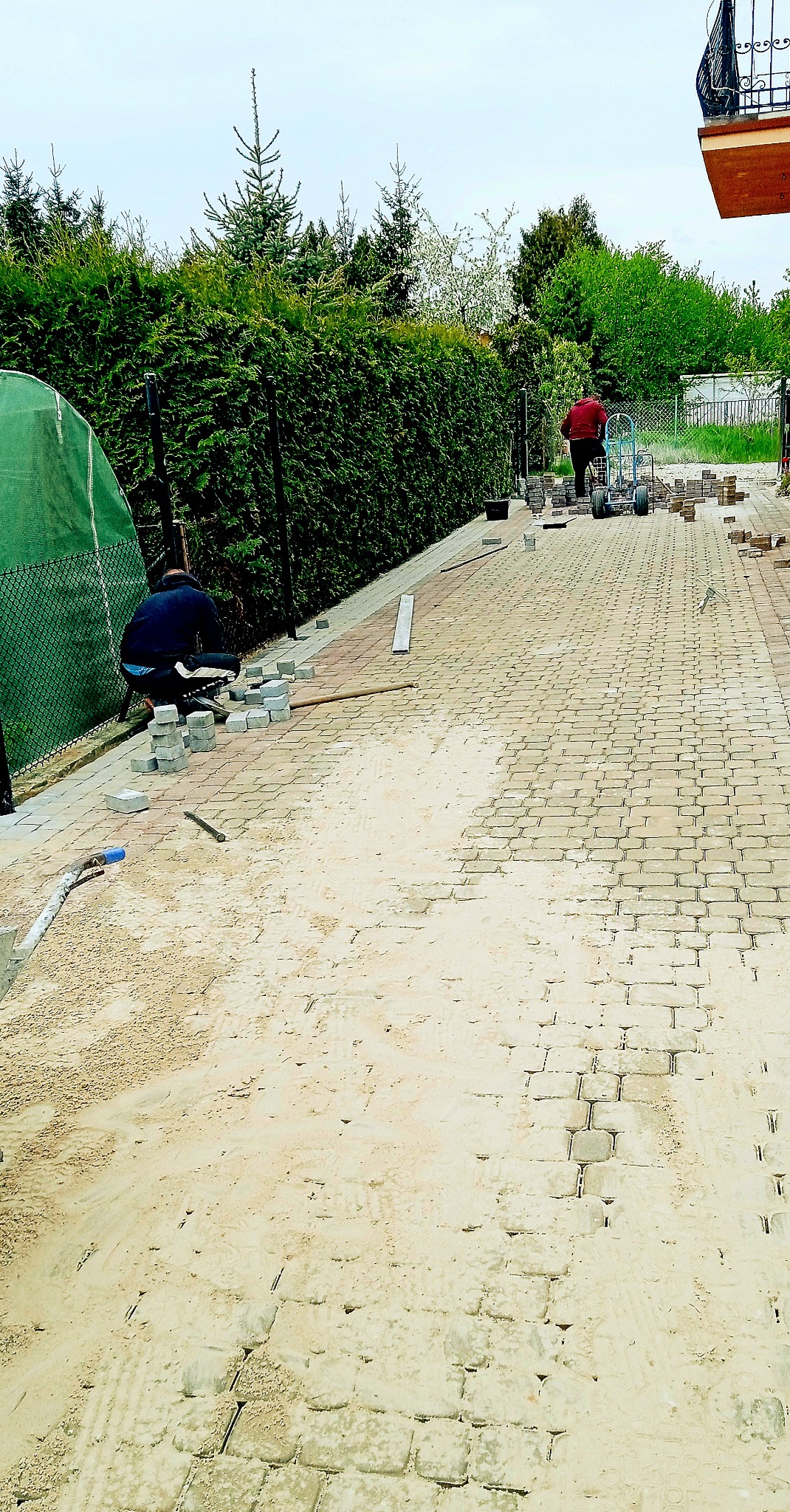 Dwóch pracowników podczas układania kostki brukowej na podjeździe; widoczny piasek, ułożona część nawierzchni z kostki, narzędzia i taczka z materiałem.