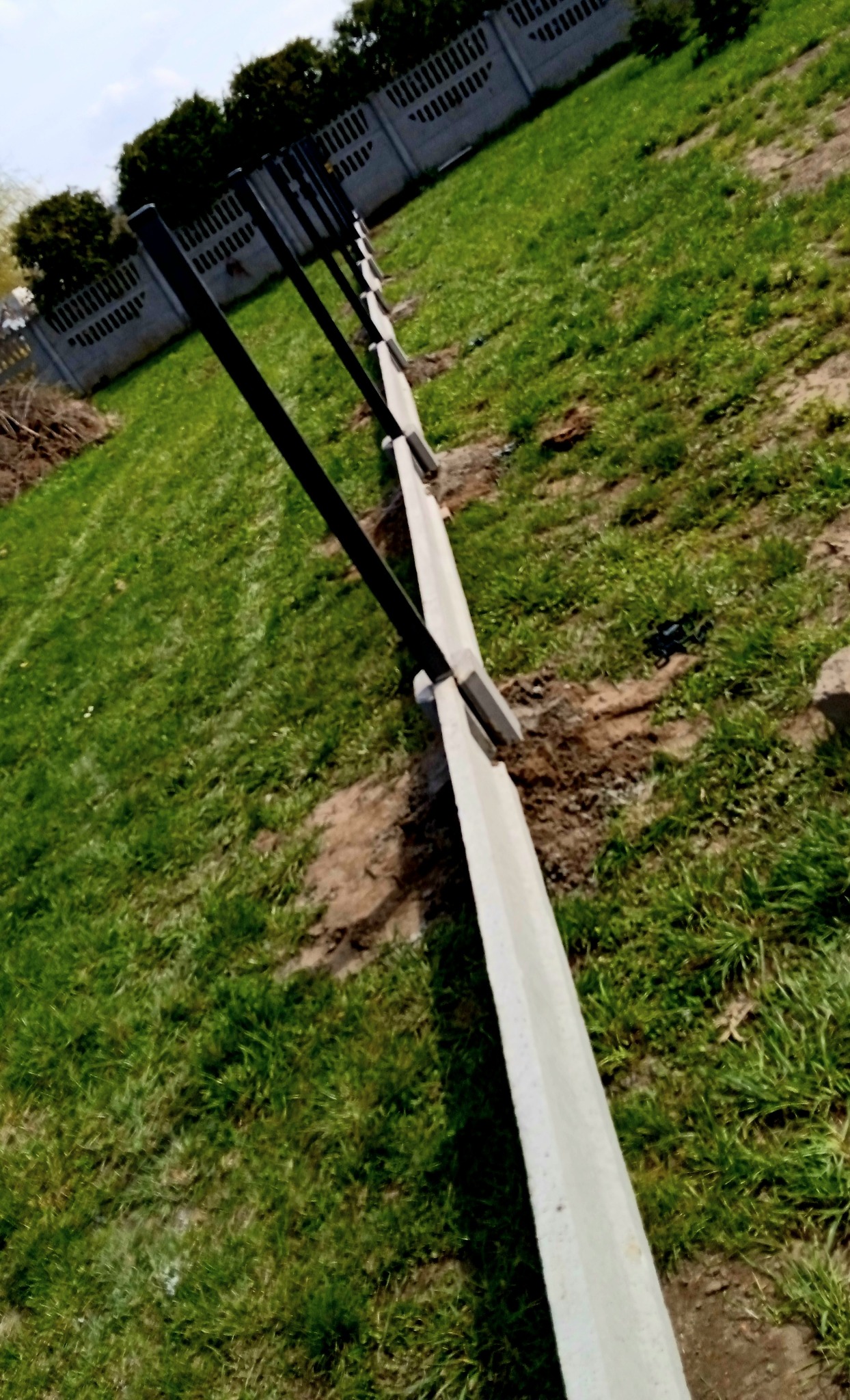 Wkopane słupki ogrodzeniowe z betonową podmurówką w trakcie montażu na trawiastym terenie, widoczne narzędzia i fragment istniejącego ogrodzenia w tle.