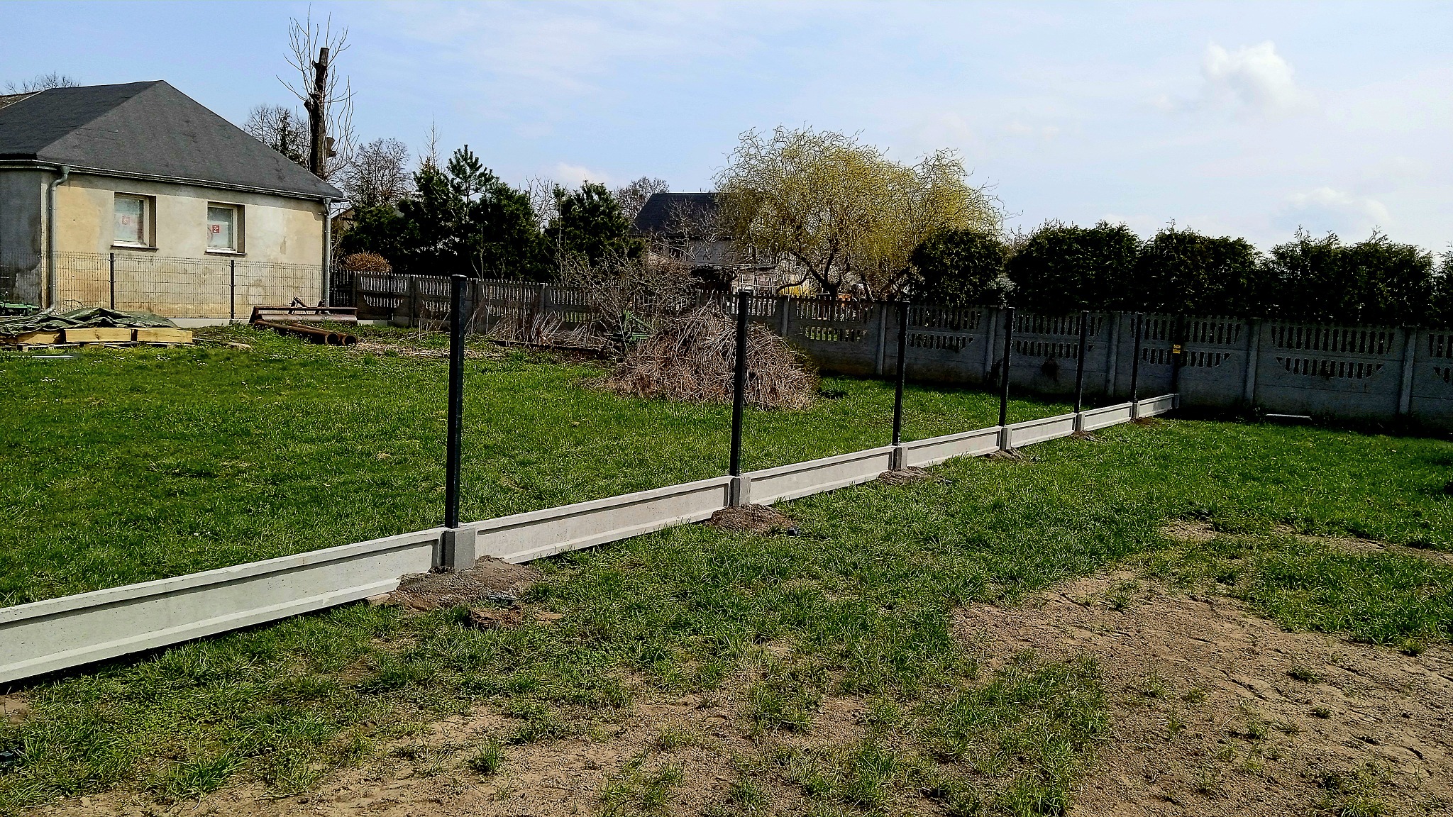 Wkopane betonowe podmurówki i słupki ogrodzeniowe, przygotowanie do montażu ogrodzenia panelowego na nierównym terenie.