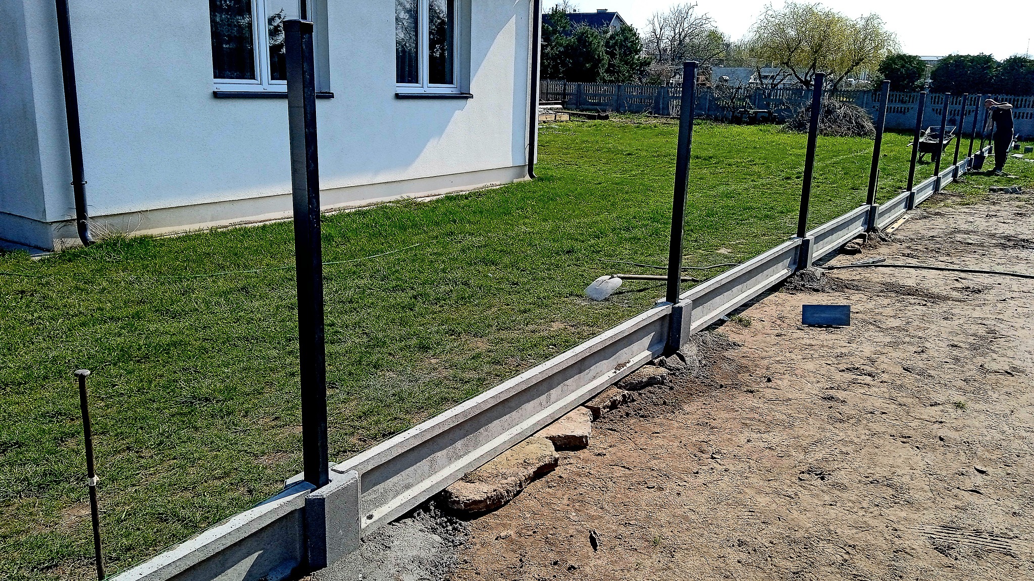 Wykonywanie fundamentu pod ogrodzenie z prefabrykowanych elementów betonowych, widoczne czarne słupki, w tle trawnik i budynek.