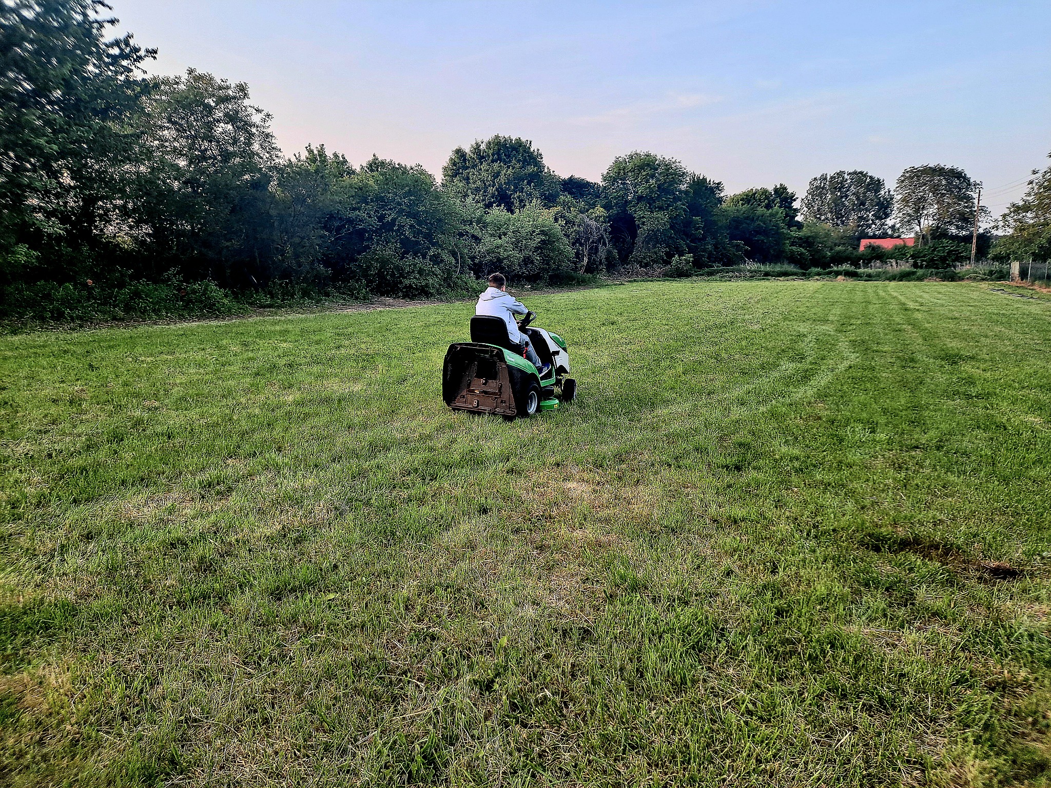 Mężczyzna w jasnej koszuli kosi trawę na rozległym, zielonym trawniku za pomocą traktorka kosiarki, na tle drzew i błękitnego nieba.
