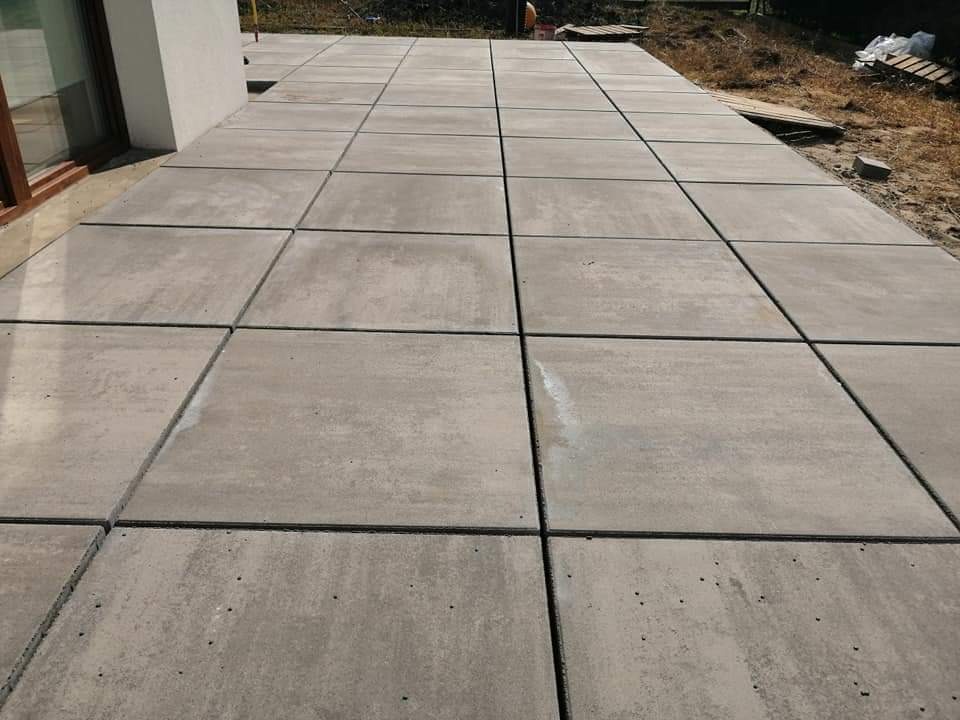 Ułożona nawierzchnia z dużych, kwadratowych płyt betonowych w odcieniach szarości, widoczne spoiny między płytami, zarys budynku w tle.