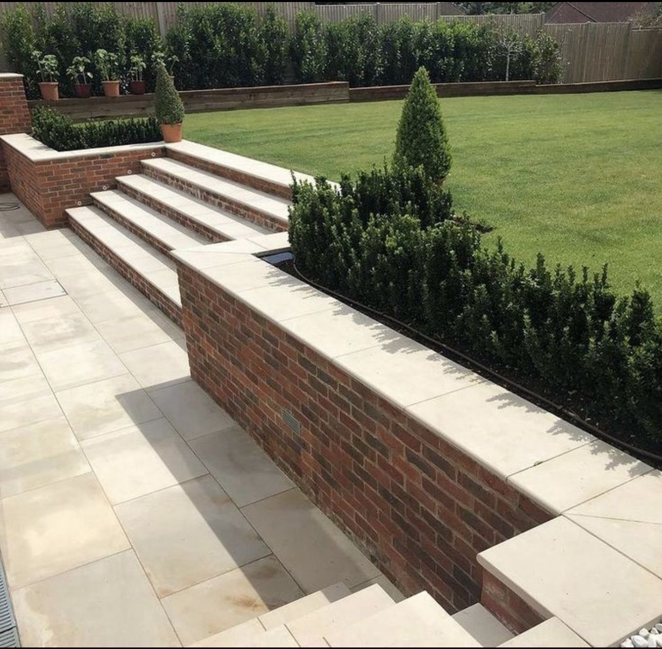 Ogród z tarasem i schodami wyłożonymi jasną kostką brukową, ceglana ścianka z wbudowanymi rabatami, trawnik z równo przyciętą trawą i żywopłotem w tle.