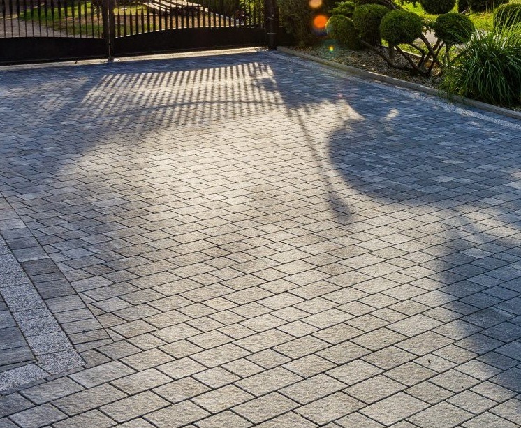 Ułożona kostka brukowa w odcieniach szarości, z widocznym cieniem bramy wjazdowej i fragmentem ogrodzenia panelowego w tle.