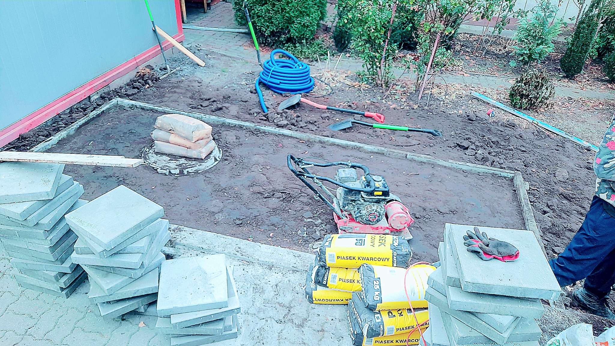 Przygotowanie terenu pod ułożenie kostki brukowej: widoczne obramowanie, ubijarka, worki z piaskiem kwarcowym, stos kostki, narzędzia i wąż ogrodowy.