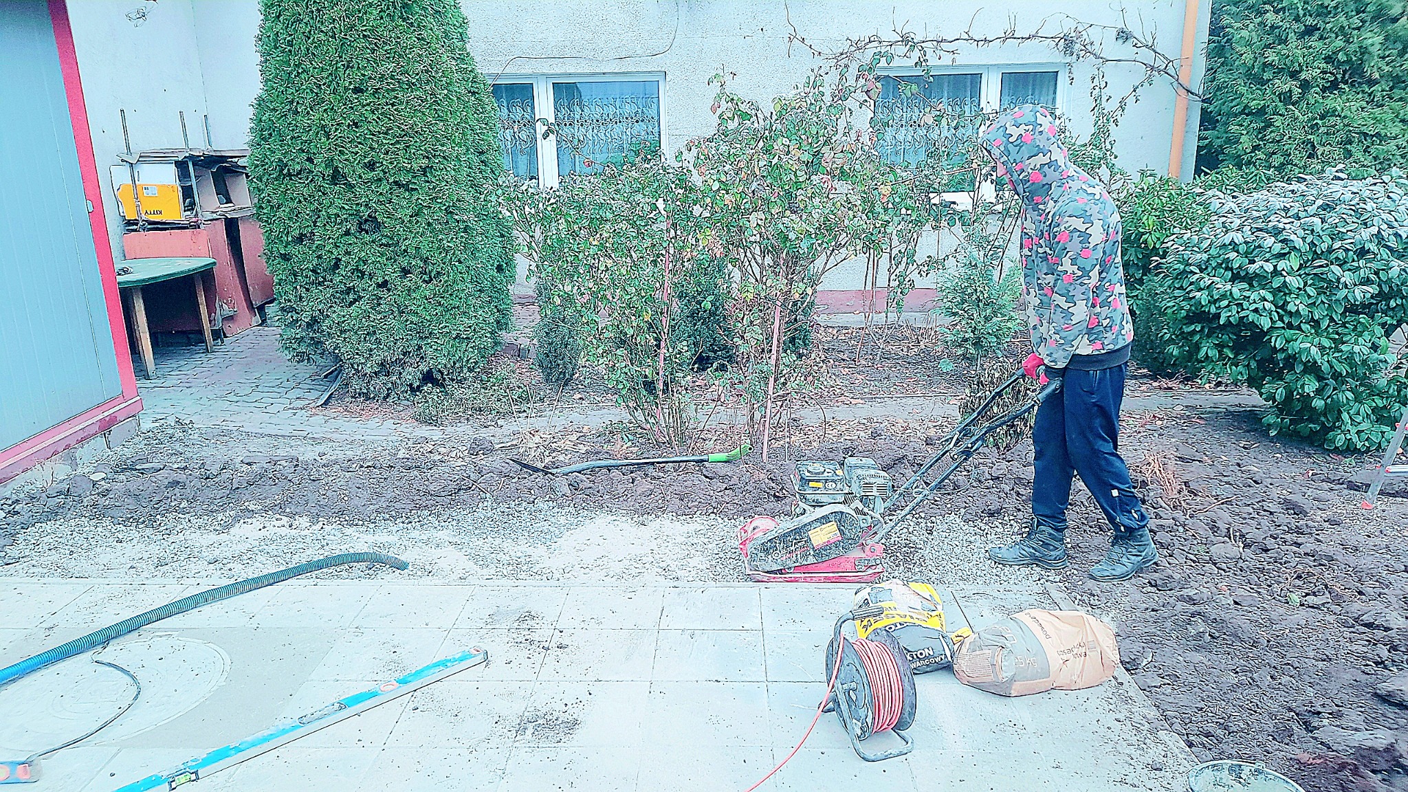 Prace przygotowawcze do ułożenia kostki brukowej: mężczyzna w kamuflażowej bluzie zagęszcza podłoże ubijarką mechaniczną. Na pierwszym planie poziomica, zwinięty kabel i worek z piaskiem. W tle...