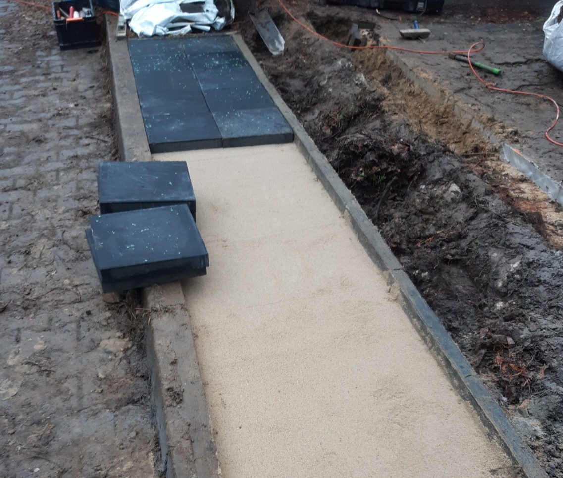 Przygotowanie podłoża z piasku pod ułożenie czarnych, kwadratowych płyt chodnikowych, widoczne narzędzia i wykop w ziemi.