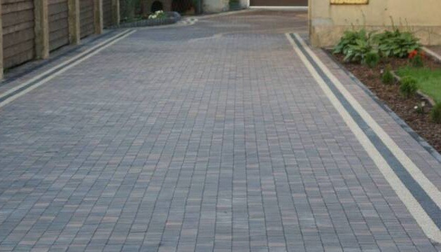 Ułożona kostka brukowa w odcieniach szarości, z dekoracyjnymi pasami po bokach, obok rabata z zielenią i drewniane ogrodzenie.