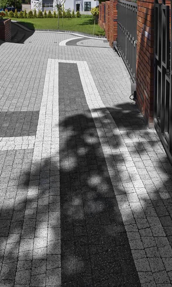 Ułożona nawierzchnia z kostki brukowej w odcieniach szarości z białymi i czarnymi akcentami, tworząca wzór geometryczny. Widoczny fragment ogrodzenia z cegły i metalu oraz cień drzewa.