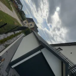 Nowoczesny garaż blaszany z bramą segmentową w kolorze antracytowym, na tle domu jednorodzinnego i pochmurnego nieba. Widok z dołu, podkreślający solidną konstrukcję.